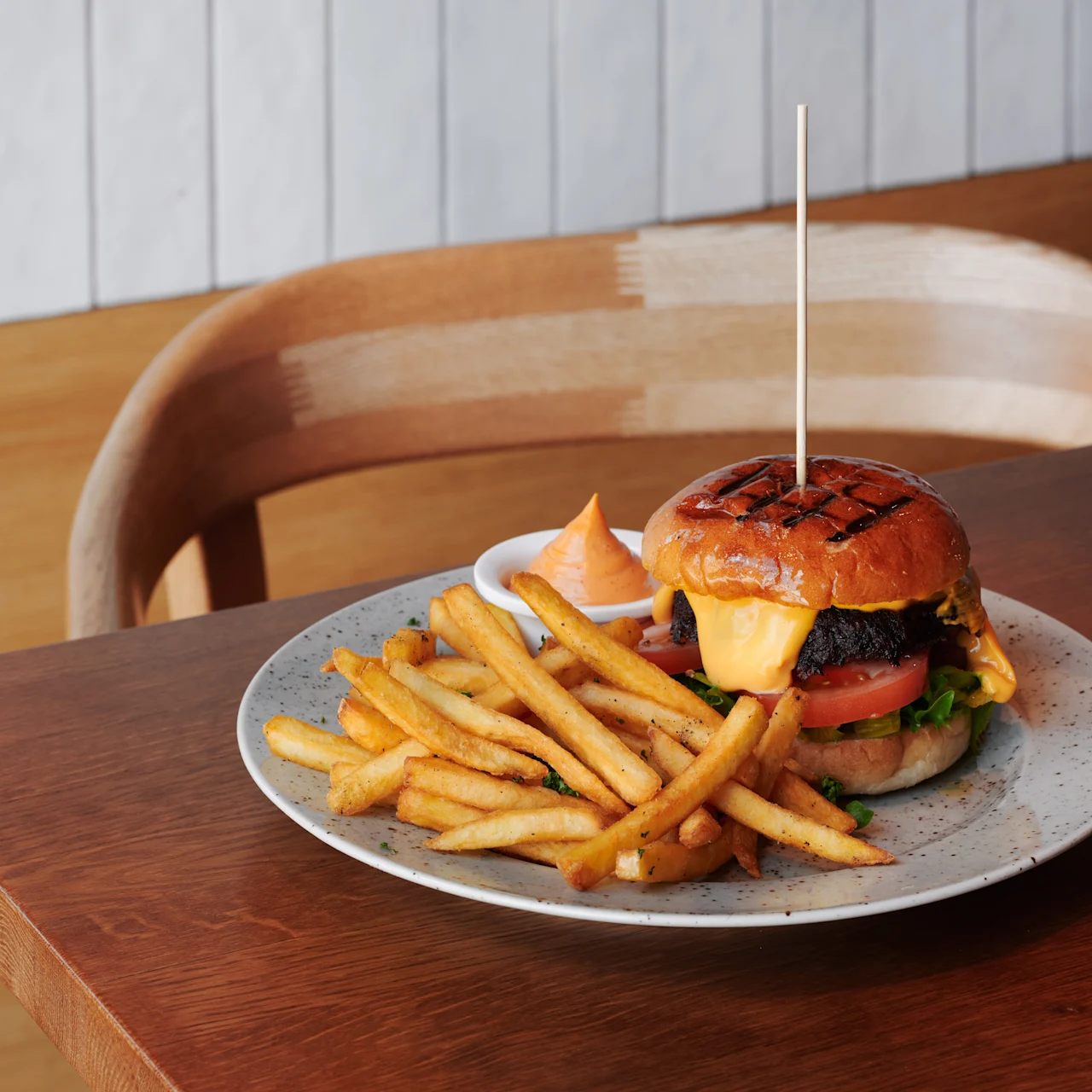 A cheeseburger with lettuce, tomato, and beef patty, served on a plate alongside golden French fries and sauce, sits on a wooden table in a restaurant setting.