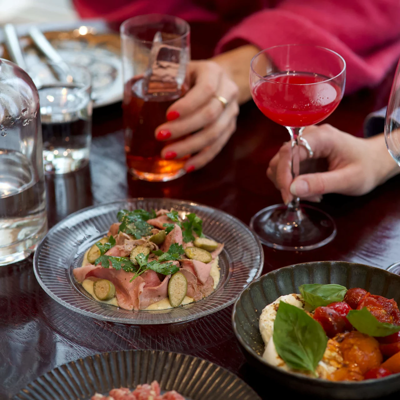 To hender som holder drikke henger over et mørkt trebord med elegante tallerkener med gourmetmat, inkludert spekemat og en tomat- og basilikumrett, i en restaurantsetting.