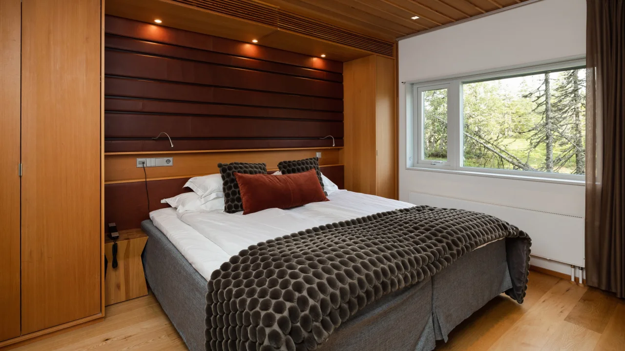 A neatly made bed against a wooden headboard, in a room with modern wooden interiors and a view of trees outside the window.