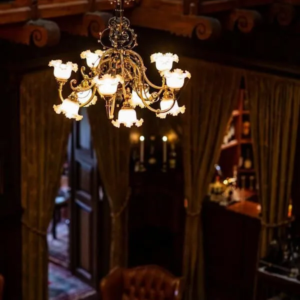 An ornate chandelier glows warmly, hanging in an elegant room with dark wood and draped curtains. In the background, an open doorway leads to a room with a patterned carpet.