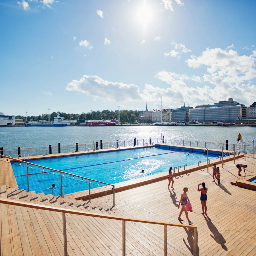 Allas Pool. Människor njuter av en solig dag vid ett utomhuspoolområde vid vattnet med en stad och ett fartyg i bakgrunden.