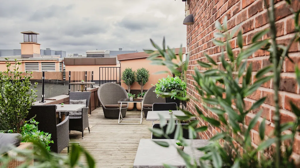 A rooftop terrace with outdoor seating, lush green plants, and a brick wall under a cloudy sky.