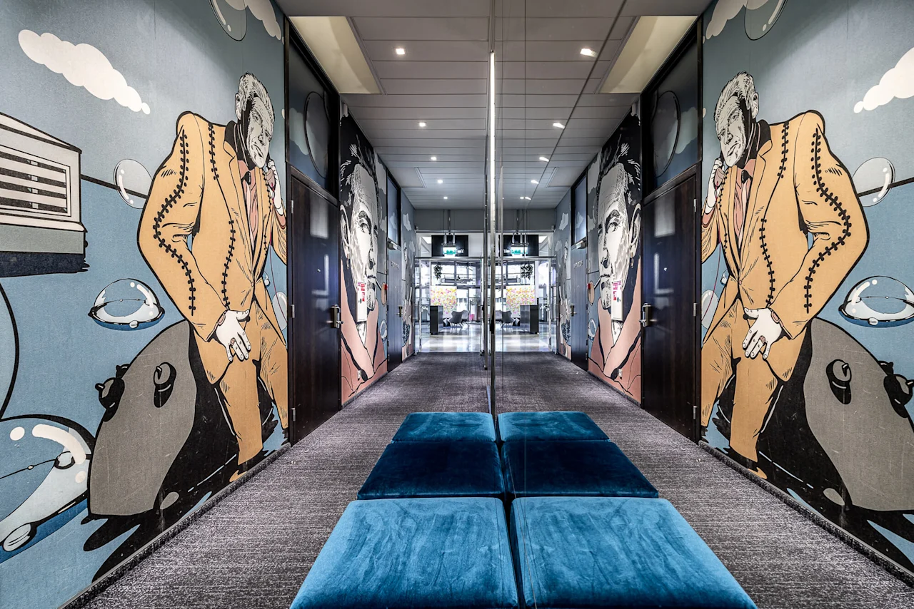 Hallway adorned with large, colorful murals of cartoon figures in suits, leading to a well-lit, open area. Blue cushioned benches sit on grey carpet flooring.