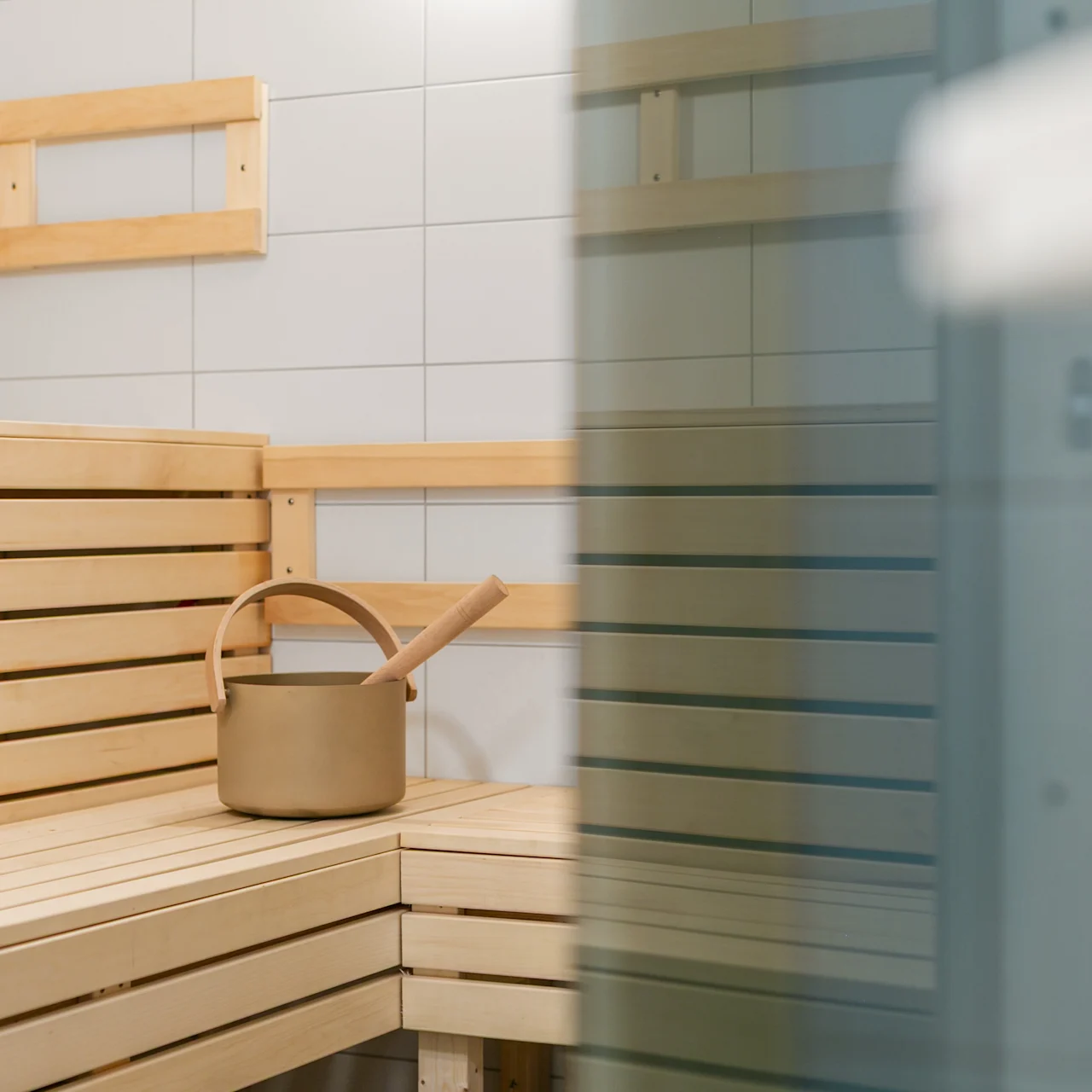 Træspand med øse på en træbænk i en sauna, omgivet af flisebelagte vægge og glasdør.