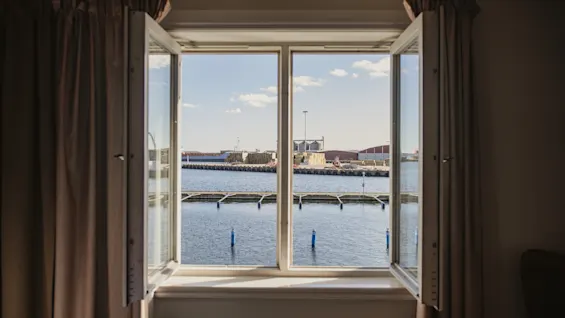En lys udsigt fra et åbent vindue med udsigt over en travl havn med roligt blåt vand og industristrukturer.