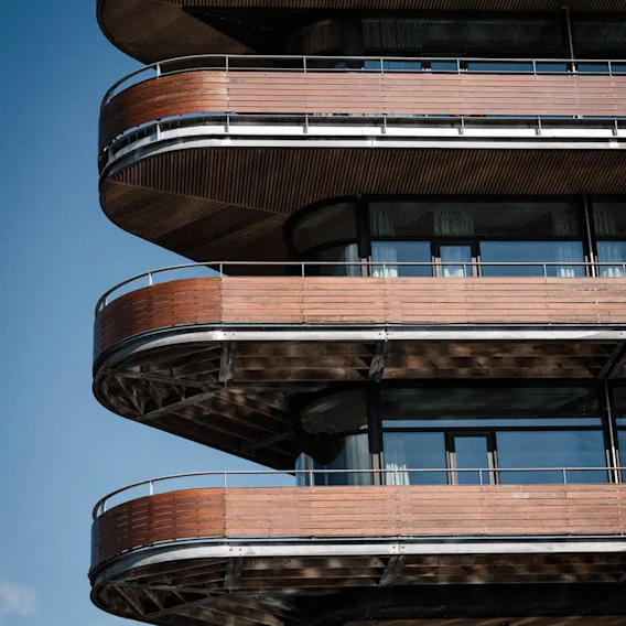 Fasadebilde av et moderne hotell med særpregede buede trealtaner mot en klar, blå himmel.