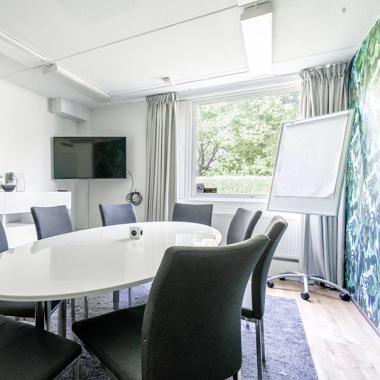 Et moderne mødelokale har et slankt hvidt ovalt bord omgivet af mørke stole. Et fladskærms-tv hænger på væggen; stort vindue tilbyder udsigt over træer.