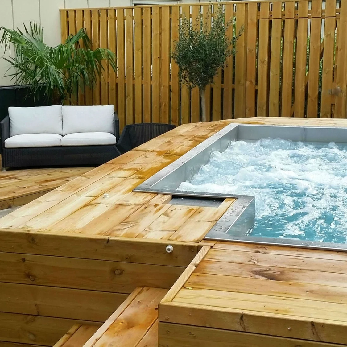 Jacuzzi, bubbling energetically, situated on a wooden deck. Surrounded by lounge chairs, a sofa, a table, and potted plants against a wooden fence.