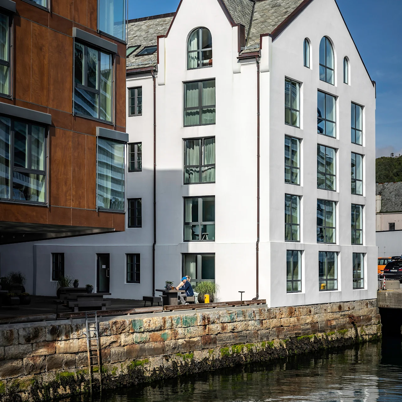 En vit byggnad vid vattnet med stora fönster står bredvid en kanal. En person i blått sitter på en bänk vid en stenbrygga, omgiven av modern arkitektur under en klar himmel.