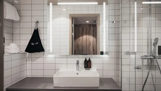 A modern bathroom with a sink, illuminated mirror, and a shower area. A bag labeled Hair dryer hangs nearby.