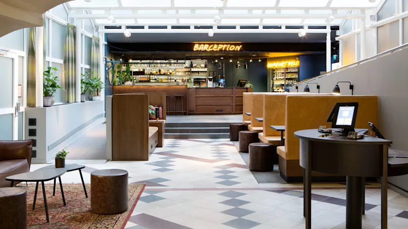 A modern hotel lobby with a bar counter labeled "BARCEPTION", seating area, tiled floor, and bright windows.