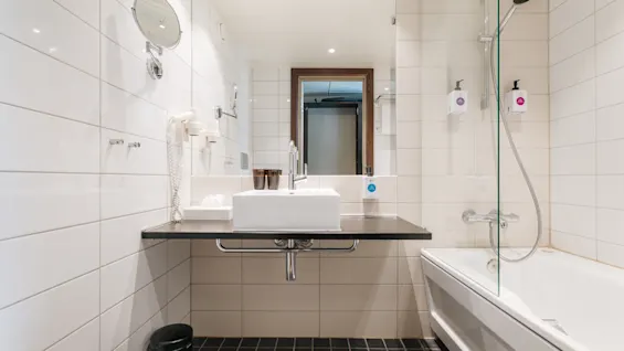 Ljust och modernt hotellbadrum med vitt kakel, handfat, stor spegel och ett badkar med dusch.