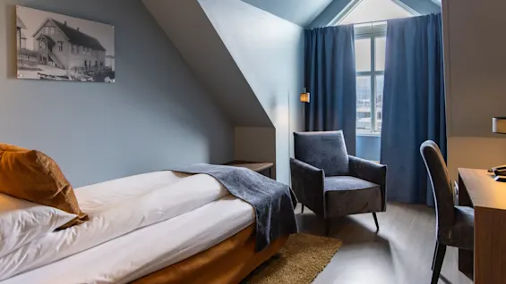 A neatly made single bed with a brown headboard stands in a blue-walled loft bedroom, accompanied by a gray armchair and desk under a pitched ceiling with a window.
