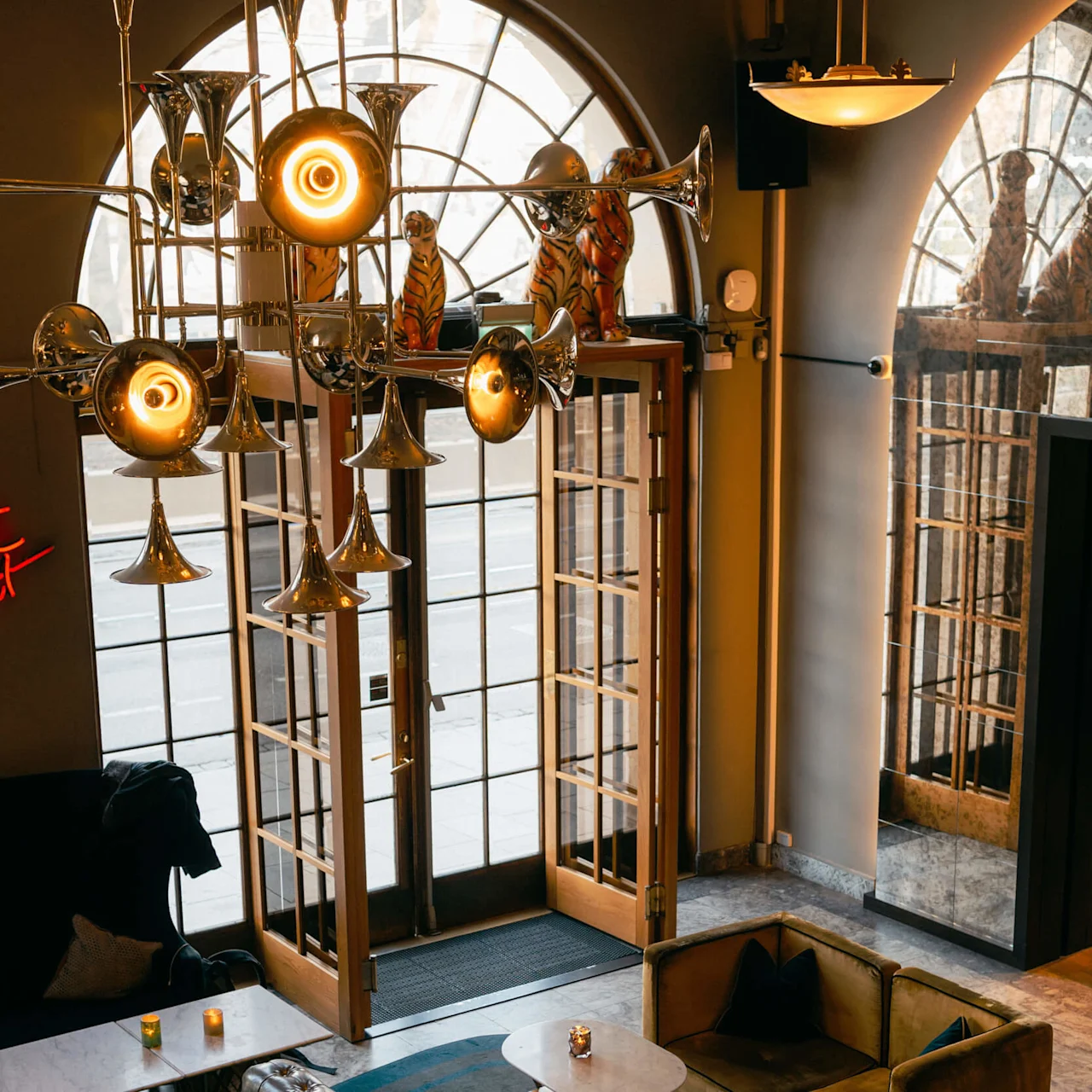 A metallic trumpet-shaped chandelier hangs from the ceiling, illuminating a cozy seating area with leather sofas and circular tables. Large windows reveal a view of the street.
