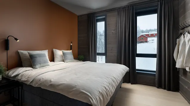 A neatly made bed with two pillows stands in a cozy bedroom, featuring brown walls and large windows overlooking a snow-covered landscape with a red house.