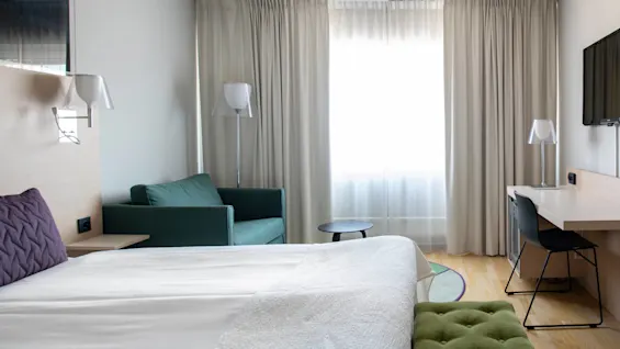 En säng med vita sängkläder står i ett hotellrum. Den vetter mot ett skrivbord med stol, en väggmonterad TV och ett fönster med beige gardiner. En grön soffa står i närheten.