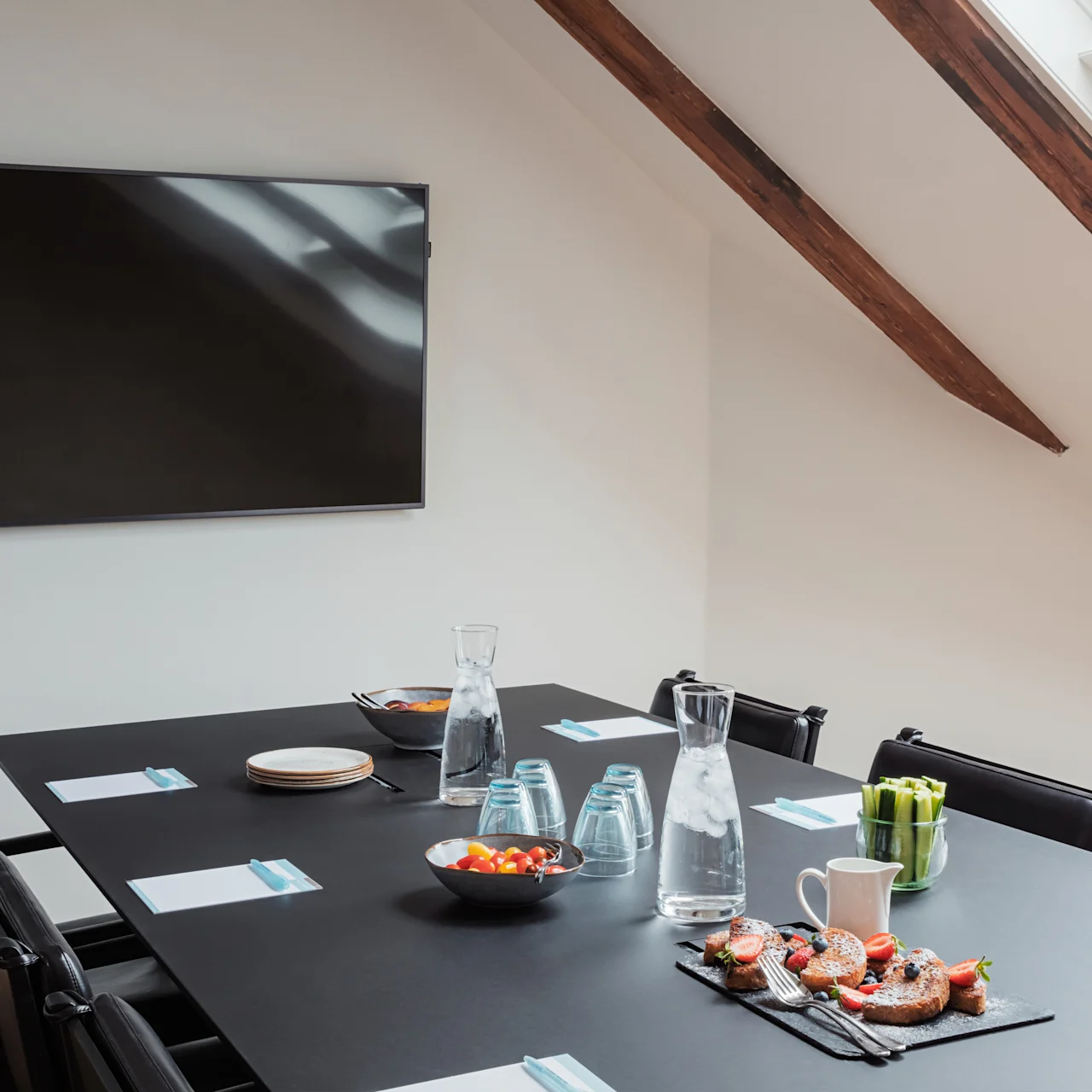 A conference table set with snacks and water jugs stands under a skylight in an attic room. Chairs surround the table, and a television is mounted on the wall.