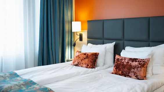 En prydligt bäddad säng på ett hotell har mjuka, sammetskuddar. En vägglampa ger mjukt ljus mot en orange vägg, med turkosa gardiner som ramar in ett fönster bredvid sängen.