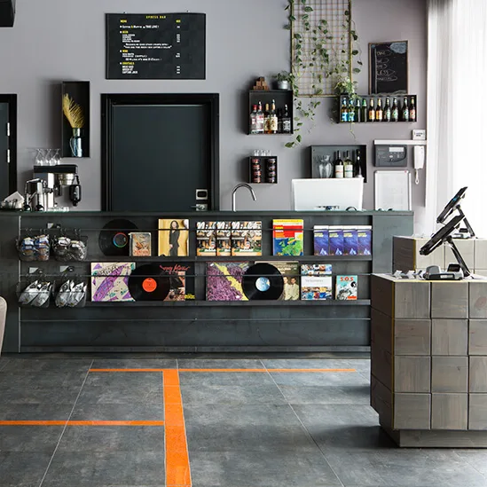 Reception desk displaying vinyl records and magazines; coffee machine and shelves with drinks and decor behind. Door marked "LIFT" with art and seating nearby.