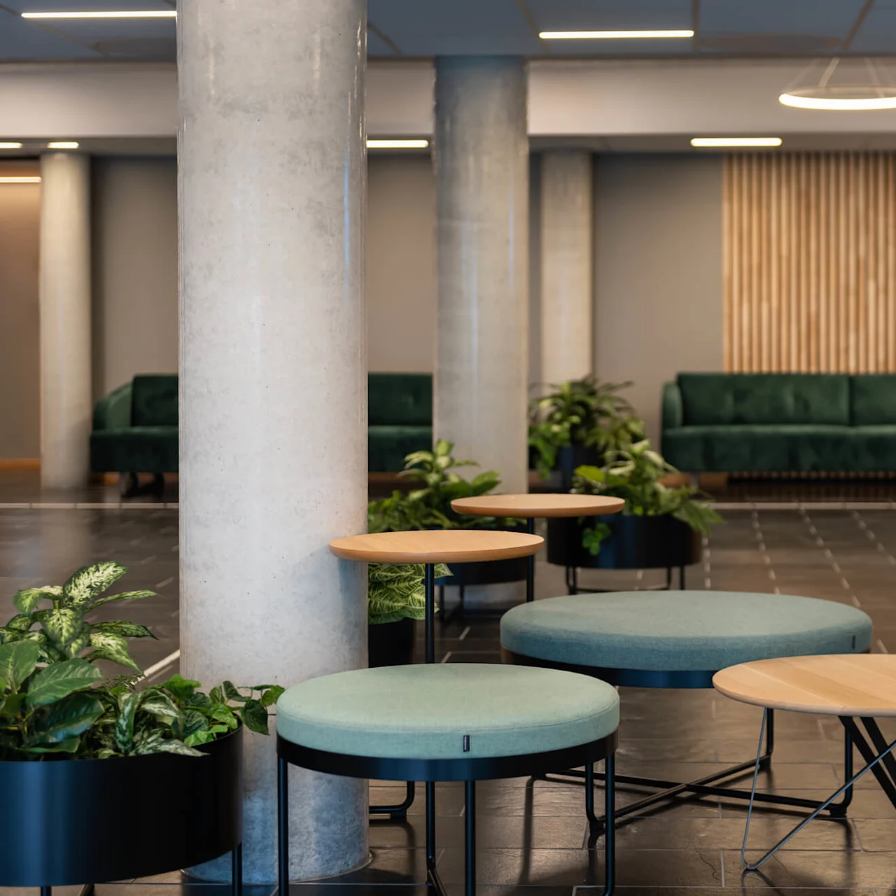 Runda bord och stoppade pallar är placerade runt krukväxter i en modern lobby med betongpelare, gröna soffor och en bakgrund av träpaneler. 