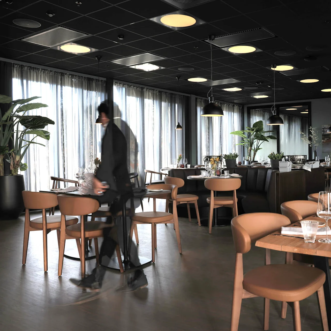 A person walks through a modern restaurant with set tables, pendant lights, and plants, suggesting preparation for service.