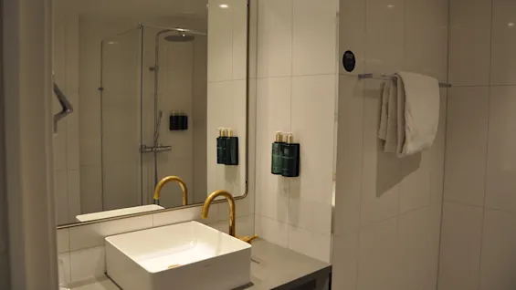 Sink with gold faucet reflected in large mirror; action: stationary; context: modern bathroom with white tiles, glass shower, and towel hanging on the wall.
