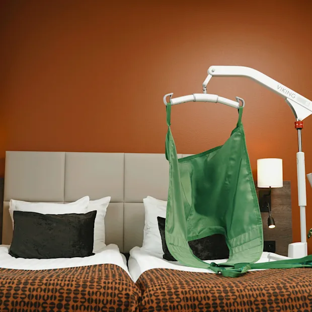 En mekanisk lyft med en grön sele svävar över två sängar i ett modernt hotellrum med orange väggar, mönstrade överkast, lampor och gardiner.