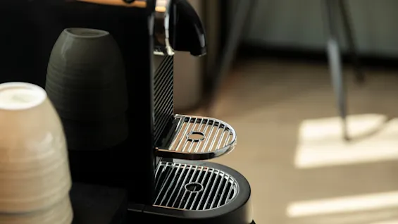 Et nærbilde av en elegant svart kaffemaskin med kromrister og to stablede keramikkkopper ved siden av, klare for en varm drikke.