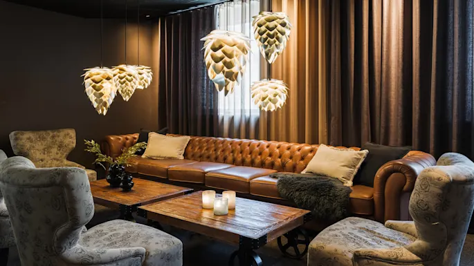 Hengende pendellamper svever over en brun skinnsofa i et koselig, svakt opplyst rom. Mønstret lenestoler og trebord med lys gir varme.