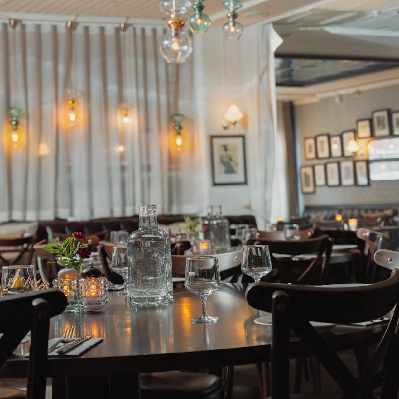 Et dækket bord med glas og en vandflaske, omringet af træstole i en svagt oplyst restaurant med varme hængende lamper og indrammede billeder på væggene.