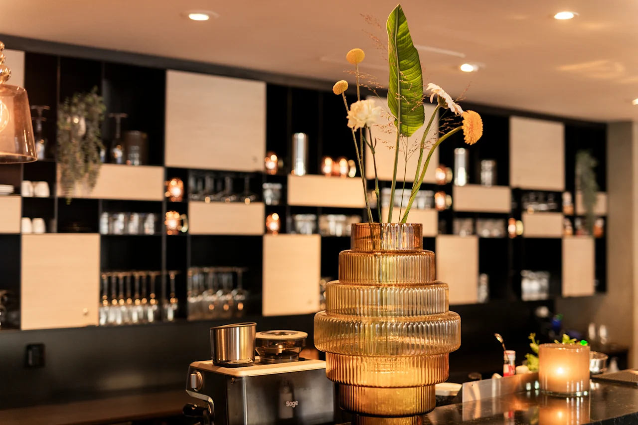 Et hyggeligt bar- eller loungeområde med et blomsterarrangement og oplyste hylder, der viser glasvarer.