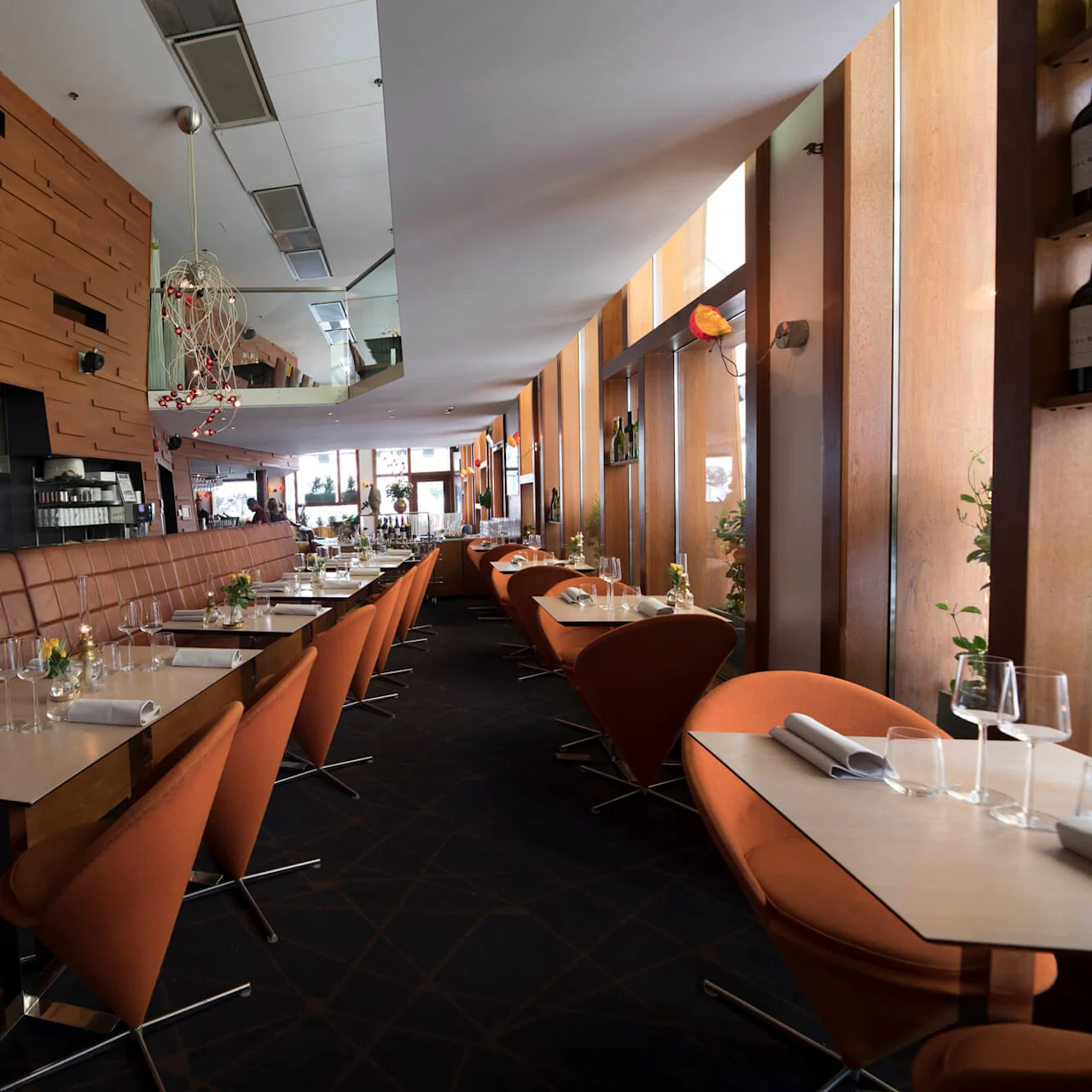 Restaurant tables with orange chairs line a modern, wooden-paneled wall. Soft lighting and decor enhance a cozy atmosphere. Glasses and napkins neatly arranged on tables. 