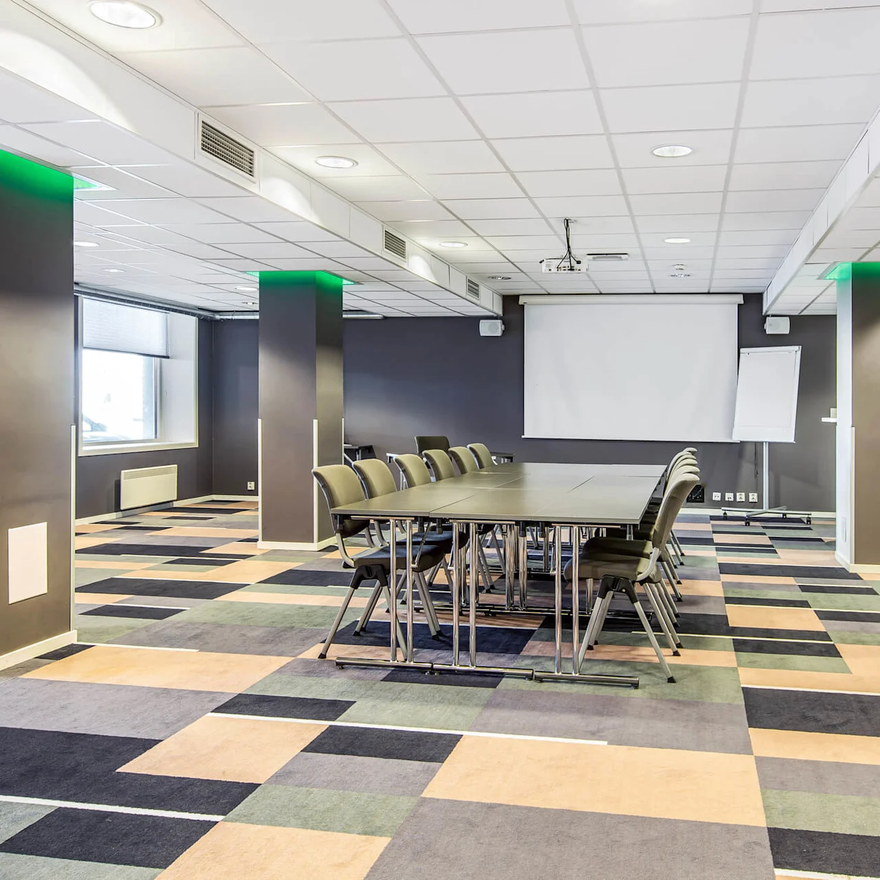 Konferansebord med stoler vender mot en projektorskjerm i et romslig, moderne rom med fargerikt geometrisk teppe og søyler opplyst av grønne lys.