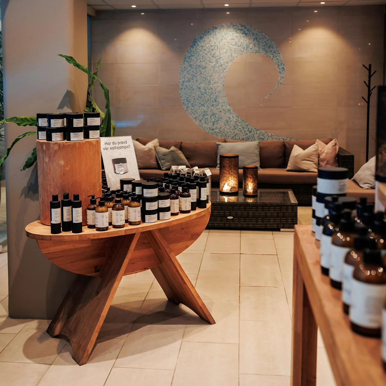 Bottles are displayed on wooden tables in a spa-like setting. A mosaic wave decorates the wall above a cozy seating area with pillows and lanterns.