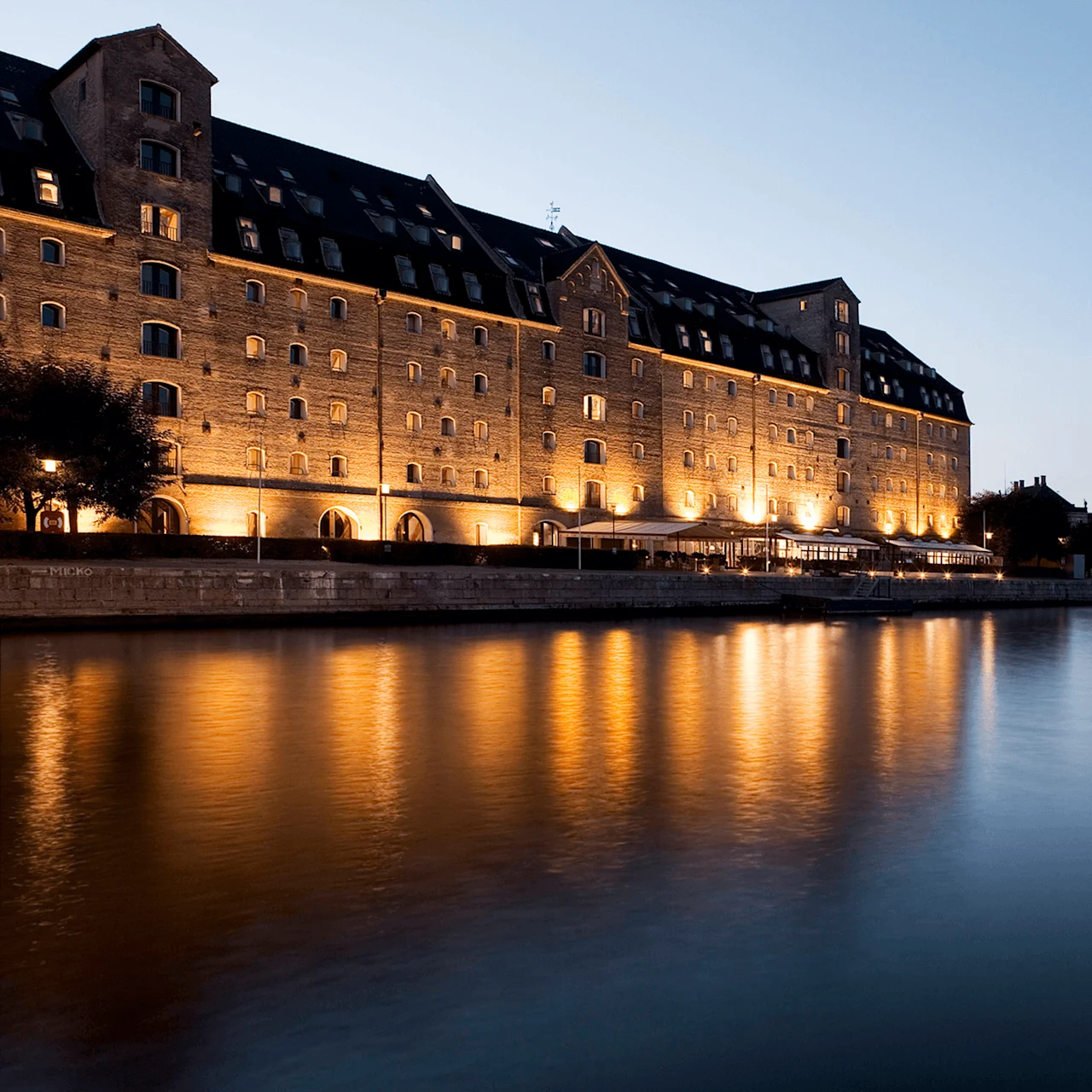 A large, illuminated brick building stands beside a calm waterfront at dusk, reflecting warm lights on the water. The sky is clear and transitioning to night.