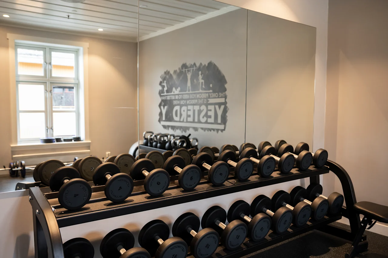 Dumbbells hviler på en rack i et godt belyst treningsrom med en speilvegg som reflekterer utstyr. Tekst på speilet: "YES I DEEP REDRM THE EKOOR" (baklengs).