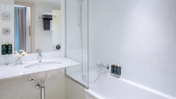 Et moderne hvidt badeværelse med en håndvask og et badekar med brusehoved. Toiletartikler er placeret på karmen. Håndklæder hænger på en stang, og blomster står på bordpladen.