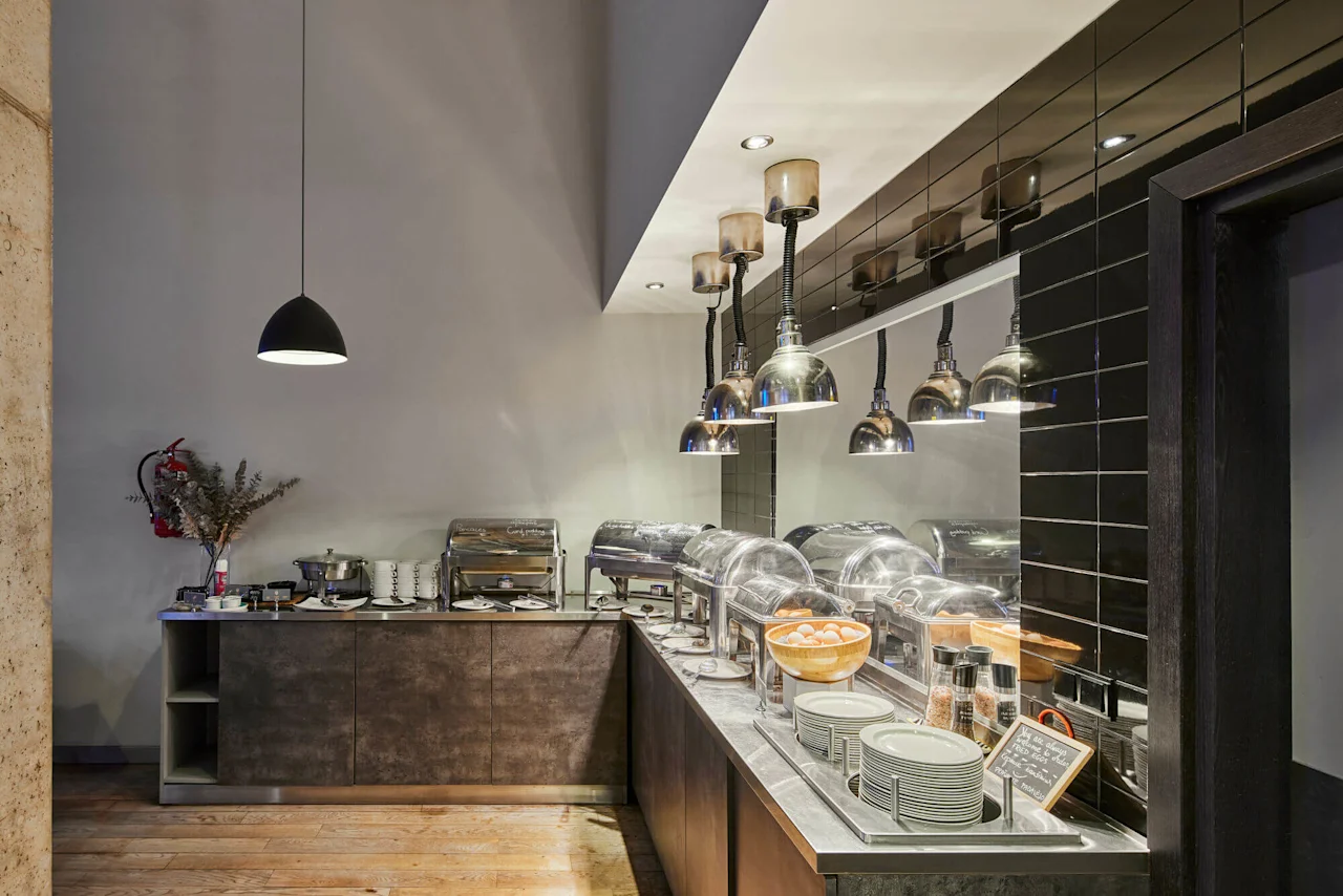Buffet setup with covered dishes on a countertop, surrounded by industrial lamps. Plates and a bowl of eggs are neatly arranged. A chalkboard sign displays: "here, we have it all."