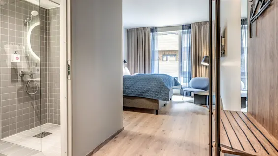 Et moderne hotelværelse har en nydeligt redt seng med blå betræk, en grå stol og trægulve. Et badeværelse med glasbruser og rundt spejl ligger ved siden af.