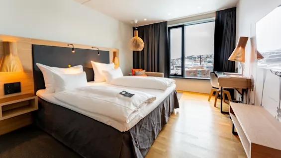 En pænt redt seng med hvidt sengetøj står i et moderne hotelværelse. Et vindue viser et sneklædt bylandskab, suppleret med stilfulde møbler i træ og blød belysning. Ingen tekst til stede.