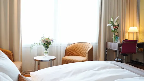 A cozy hotel room features a neatly made bed, a brown armchair beside a small table with flowers, and a desk with a lamp and another flower vase.