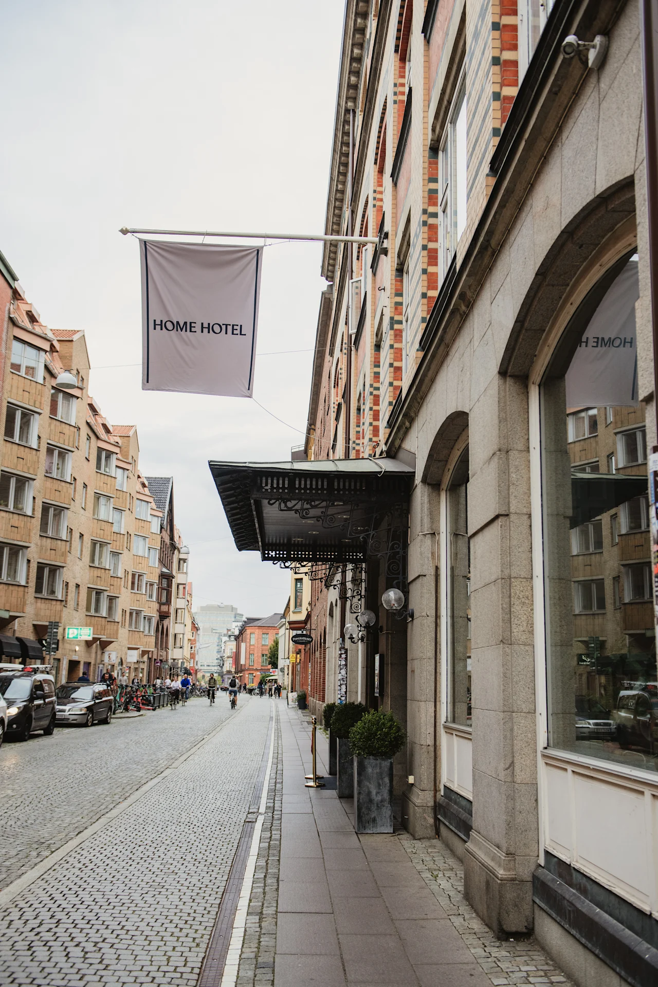 En travl europæisk gadescene med et HOME HOTEL-banner, historisk arkitektur og folk, der nyder en gåtur eller cykeltur.