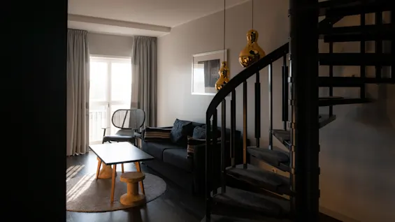 Et hyggeligt opholdsrum med en mørk sofa, sofabord og en vindeltrappe, der fører opad. Sollys strømmer ind gennem vinduet.