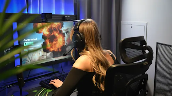 En person med hovedtelefoner spiller "Counter-Strike 2" med eksplosioner på computeren, i et rum med blå belysning og gardinbaggrund.