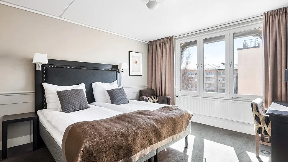 A neatly made bed takes center stage in a well-lit bedroom with windows overlooking buildings.