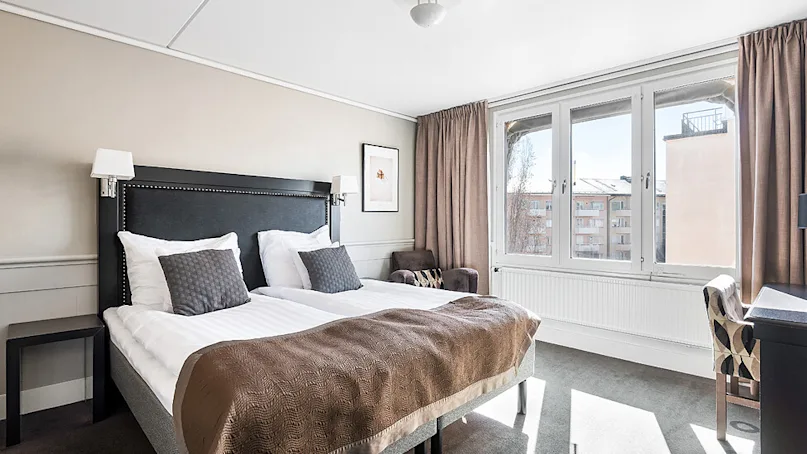 A neatly made bed takes center stage in a well-lit bedroom with windows overlooking buildings.
