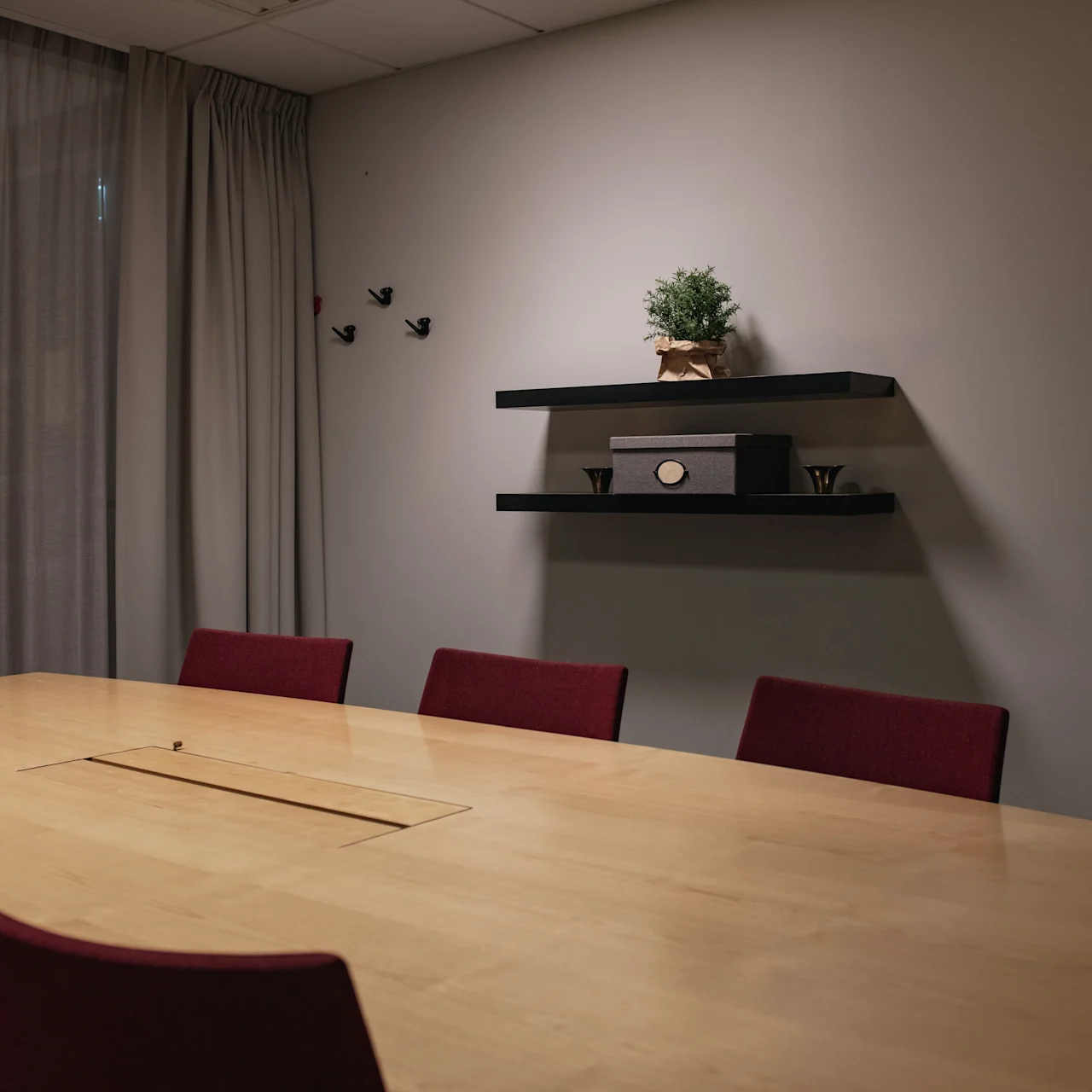 A wooden conference table surrounded by red chairs sits in a room with gray curtains. Wall shelves hold a plant and decorative items, while butterflies adorn the wall.