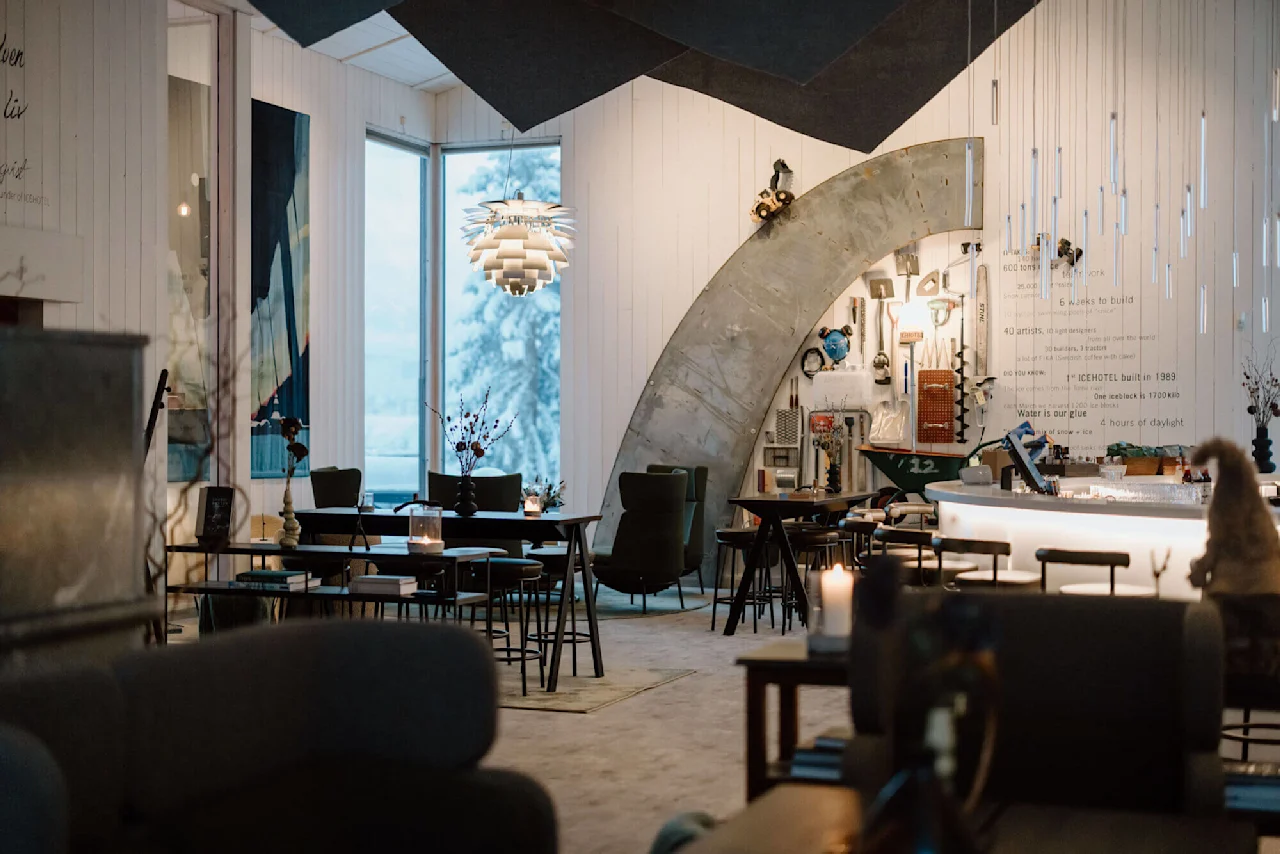 Moderne caféinteriør med eklektisk dekoration og en lysarmatur, store vinduer viser en snedækket skov. Vægtekst: "35 000 tonnes of snow and ice, 600 tons of pure work...ICEHOTEL built in 1989."