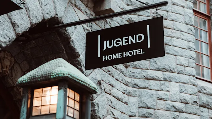 Et svart skilt med teksten "JUGEND HOME HOTEL" henger fra en steinbygning. Under sender en utsmykket gatelykt ut varmt lys, som fremhever den historiske arkitekturen.