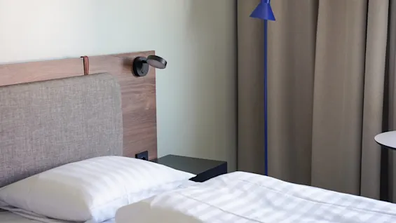 En seng med hvitt sengetøy står foran en tresengegavl. Ved siden av står en blå gulvlampe ved beige gardiner.